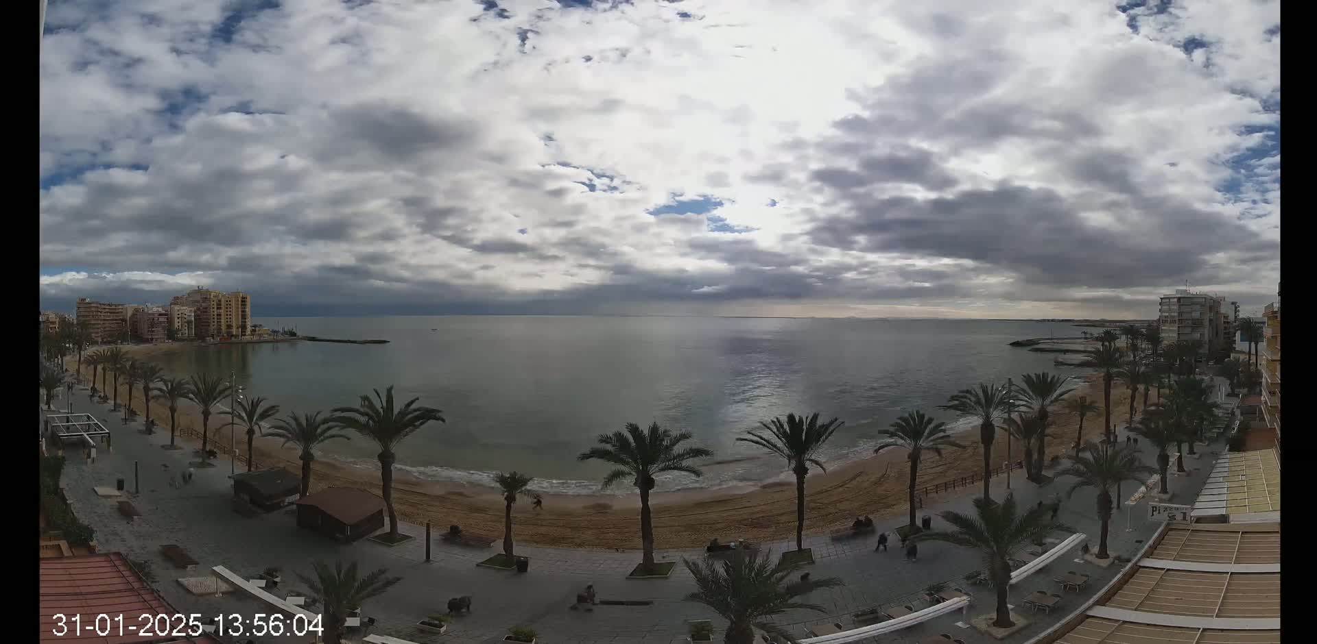 A mostly cloudy day reveals a sandy beach lined with palm trees and buildings along a calm sea.