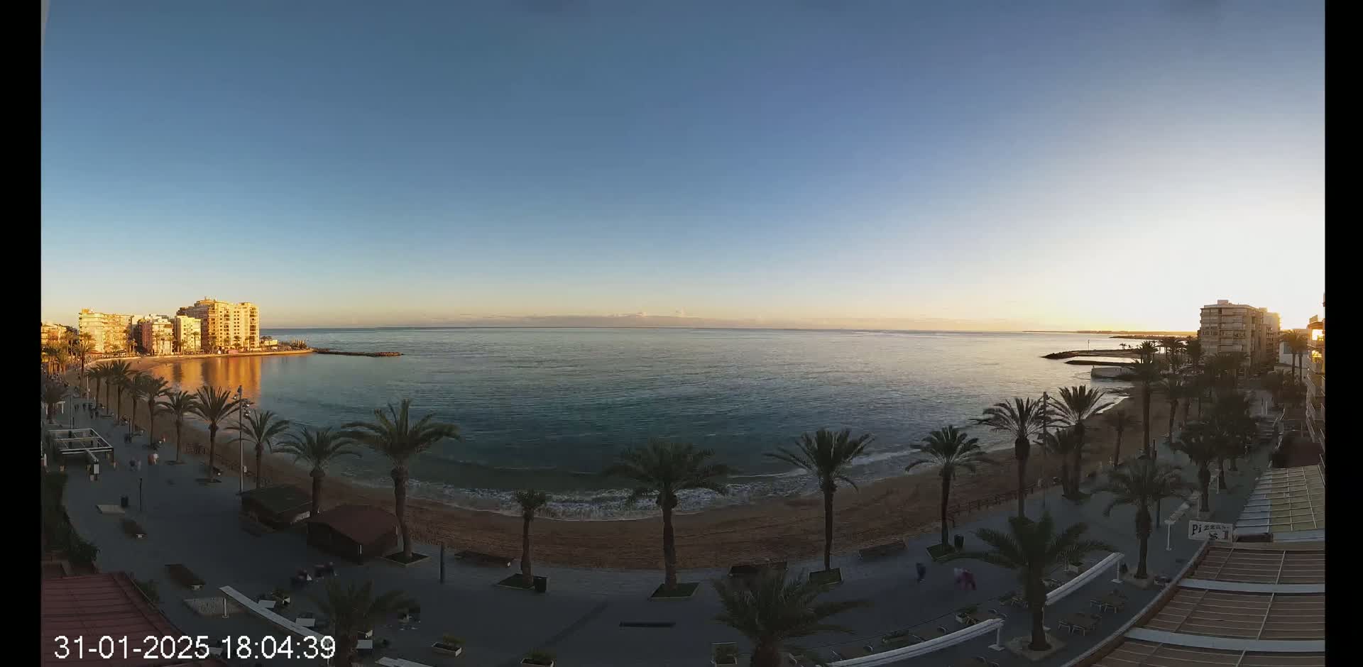 A calm ocean meets a sandy beach lined with palm trees and buildings under a clear, twilight sky.