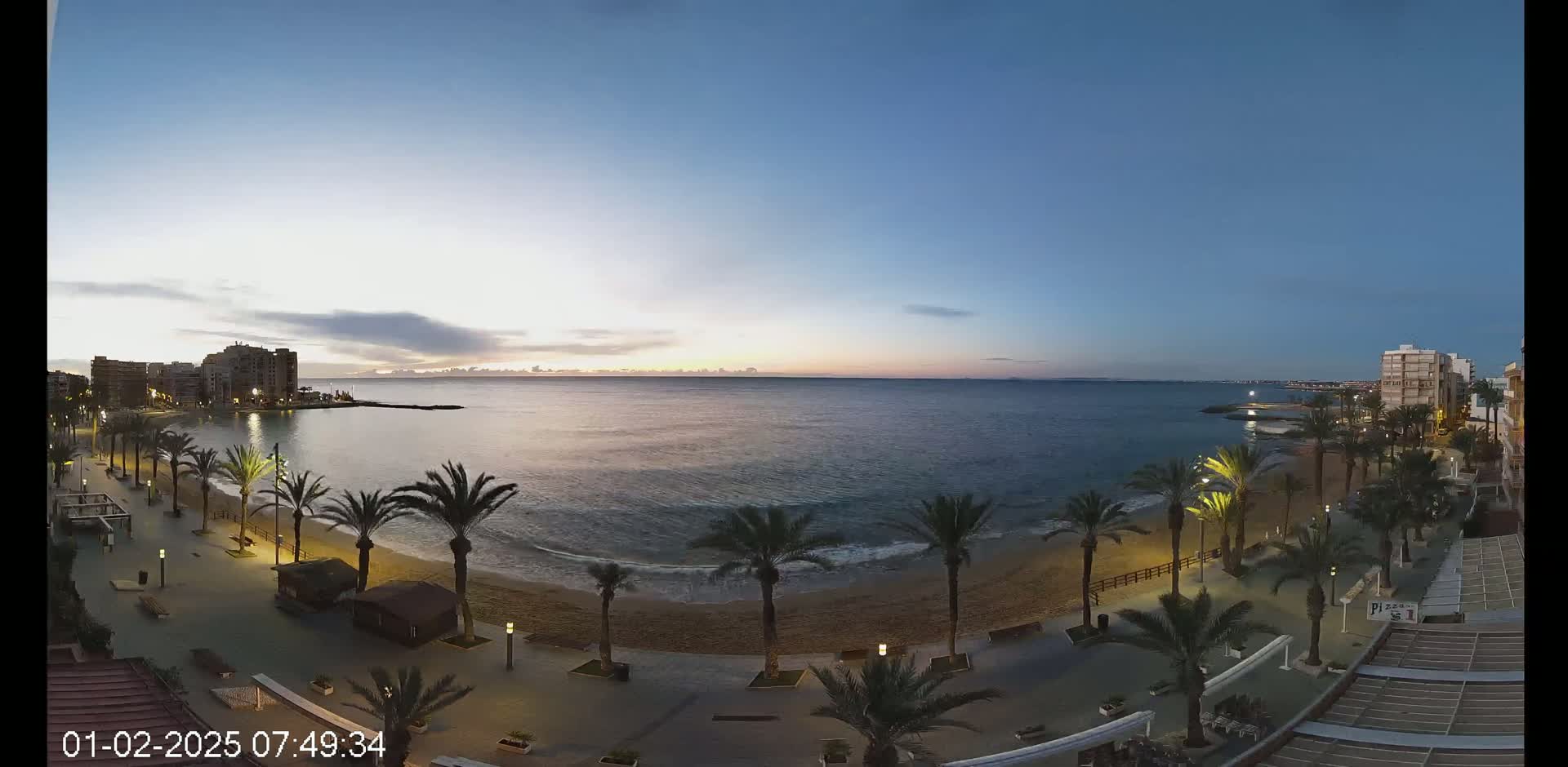 A calm ocean at sunrise is seen from above a beach promenade lined with palm trees and buildings.