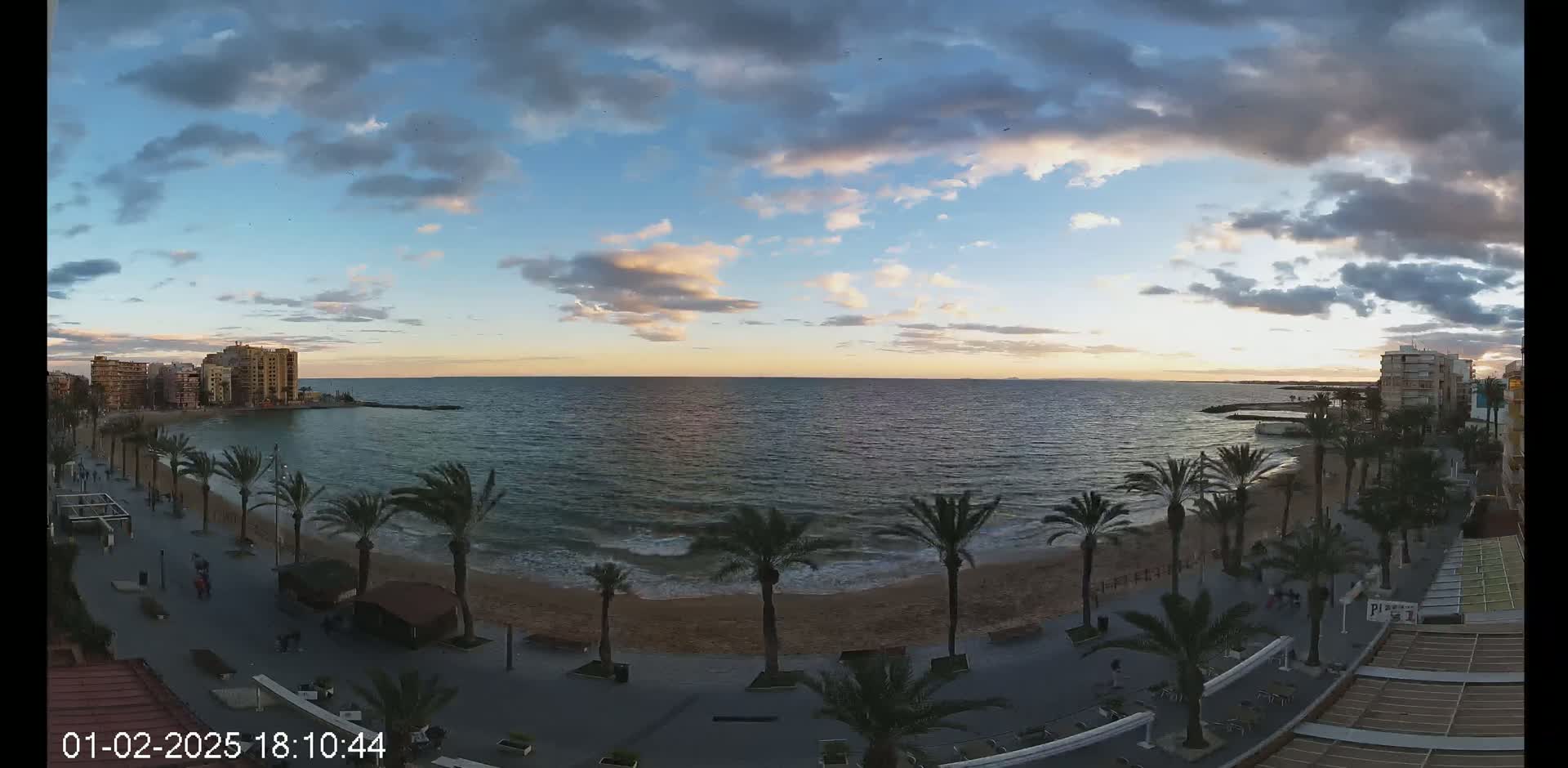A partly cloudy sunset over a calm sea with a sandy beach lined with palm trees and buildings along a promenade.
