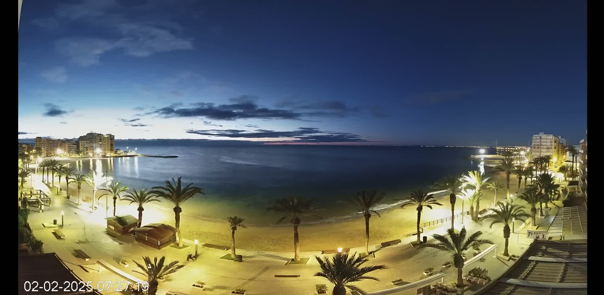 A nighttime beach scene shows a sandy shore lined with palm trees and illuminated walkways, under a mostly clear, dark blue sky.