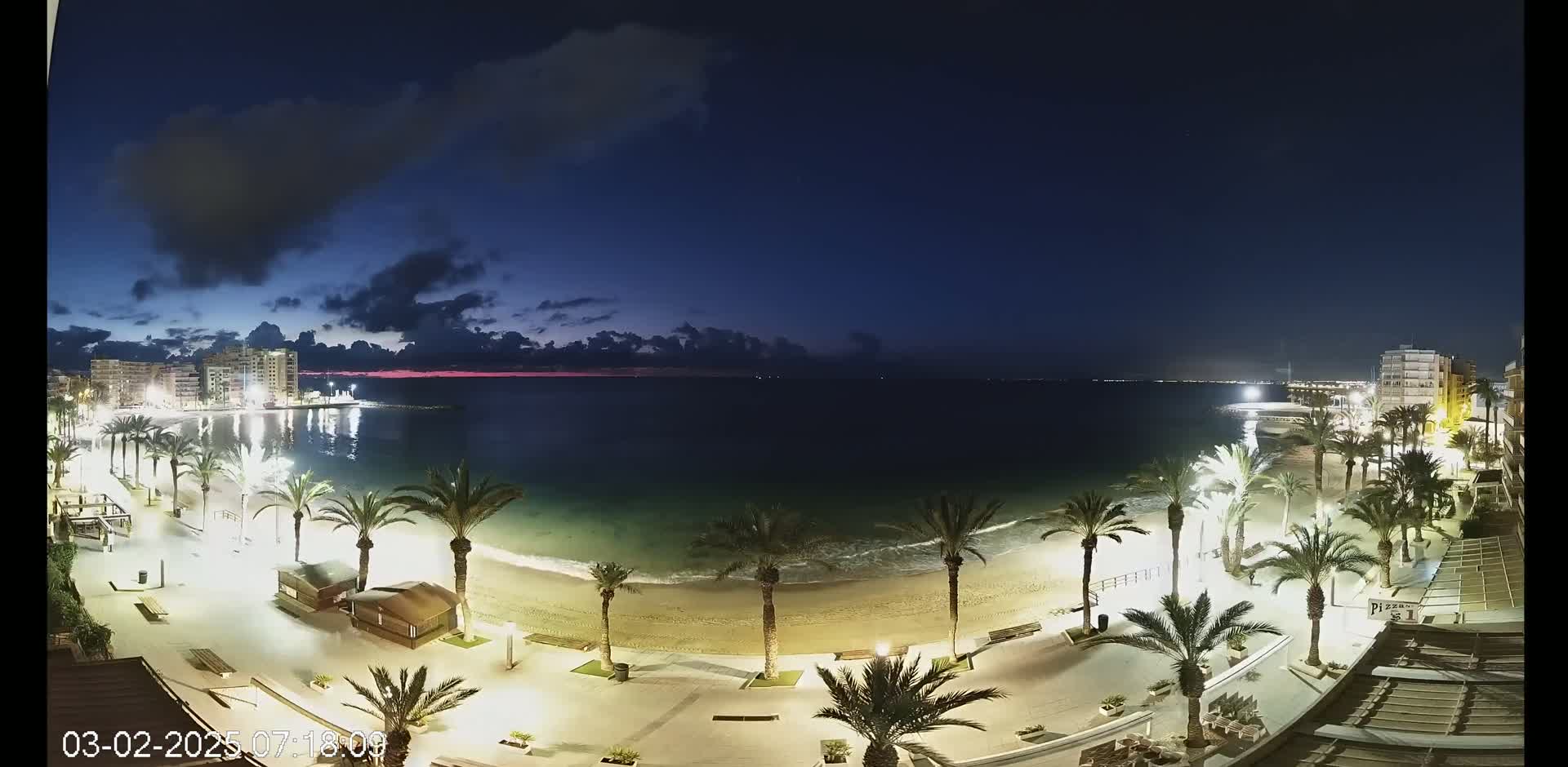 A nighttime beach scene shows a sandy shore lined with palm trees and illuminated walkways, under a mostly clear, dark blue sky with some clouds.