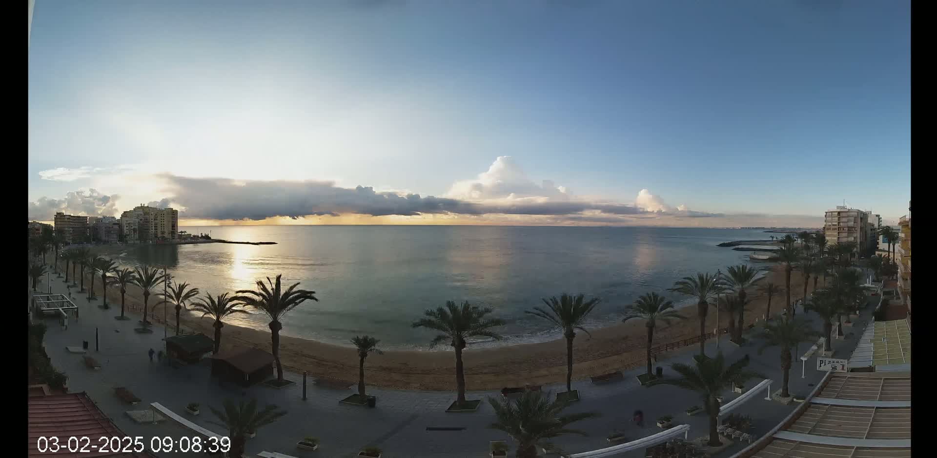 A sunrise over a calm ocean with a sandy beach lined with palm trees and buildings visible in the distance.