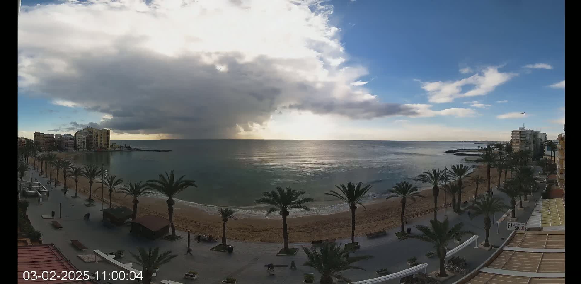 A mostly cloudy day with a dark, ominous cloud formation over a calm sea, palm trees lining a sandy beach and promenade with buildings in the background.