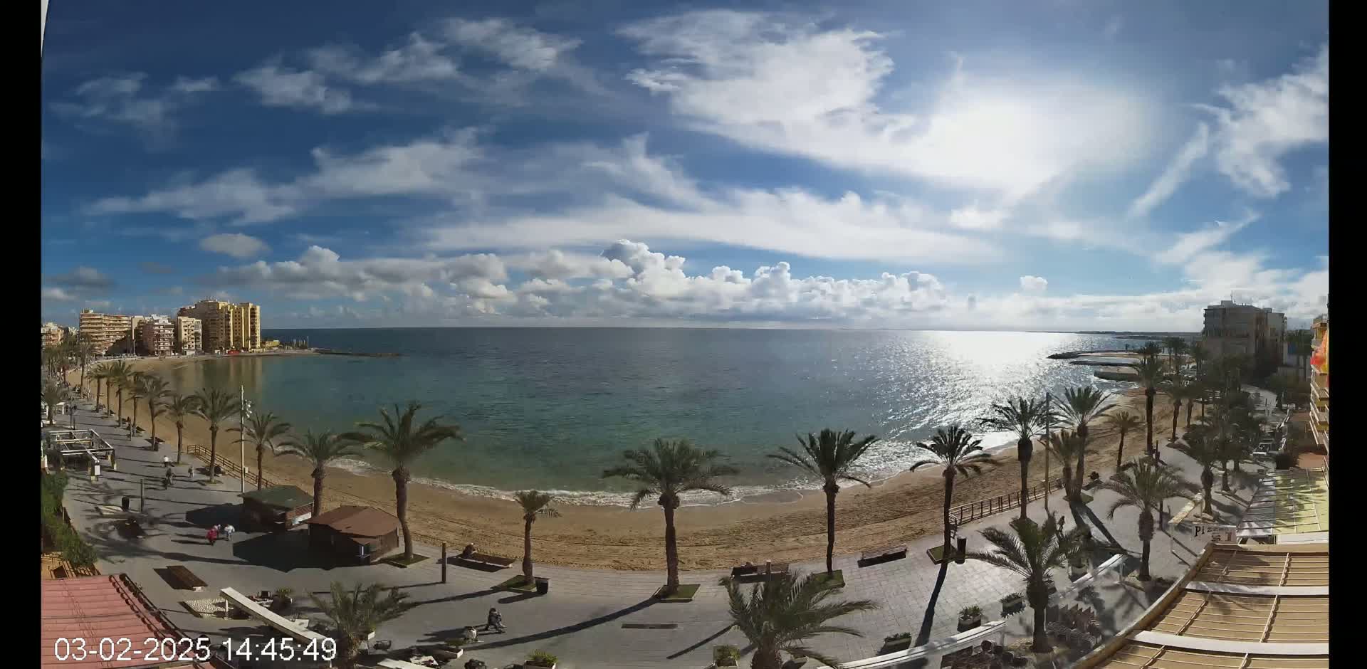 A sunny day with scattered clouds reveals a sandy beach lined with palm trees and a boardwalk, overlooking a calm sea and buildings in the distance.
