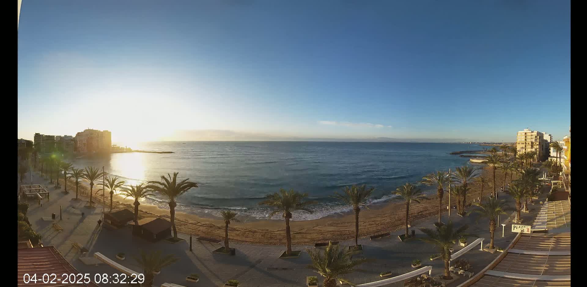 A clear sunrise illuminates a sandy beach lined with palm trees and buildings, with calm ocean waves lapping the shore.