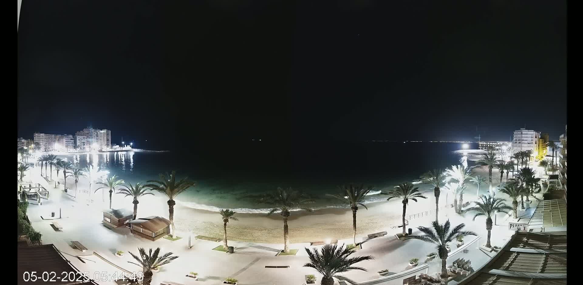 A nighttime view of a sandy beach lined with palm trees and illuminated walkways, with buildings visible in the distance across a calm body of water.