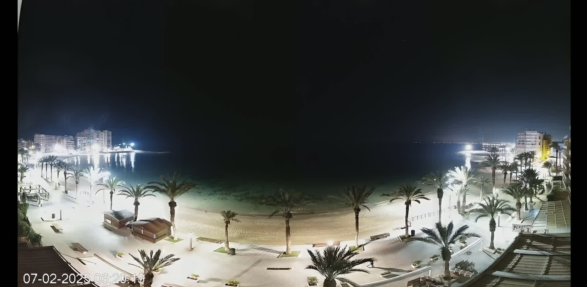 A nighttime aerial view of a sandy beach lined with palm trees and illuminated buildings along a calm body of water.