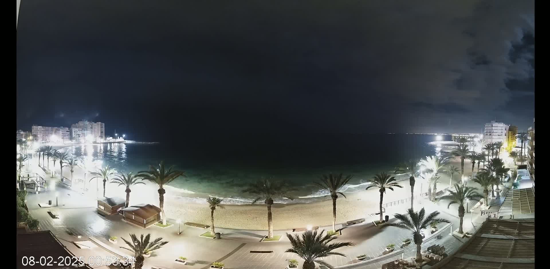 A nighttime beach scene shows a sandy shore with gentle waves, palm trees lining a paved walkway, and buildings illuminated in the distance under a partly cloudy sky.