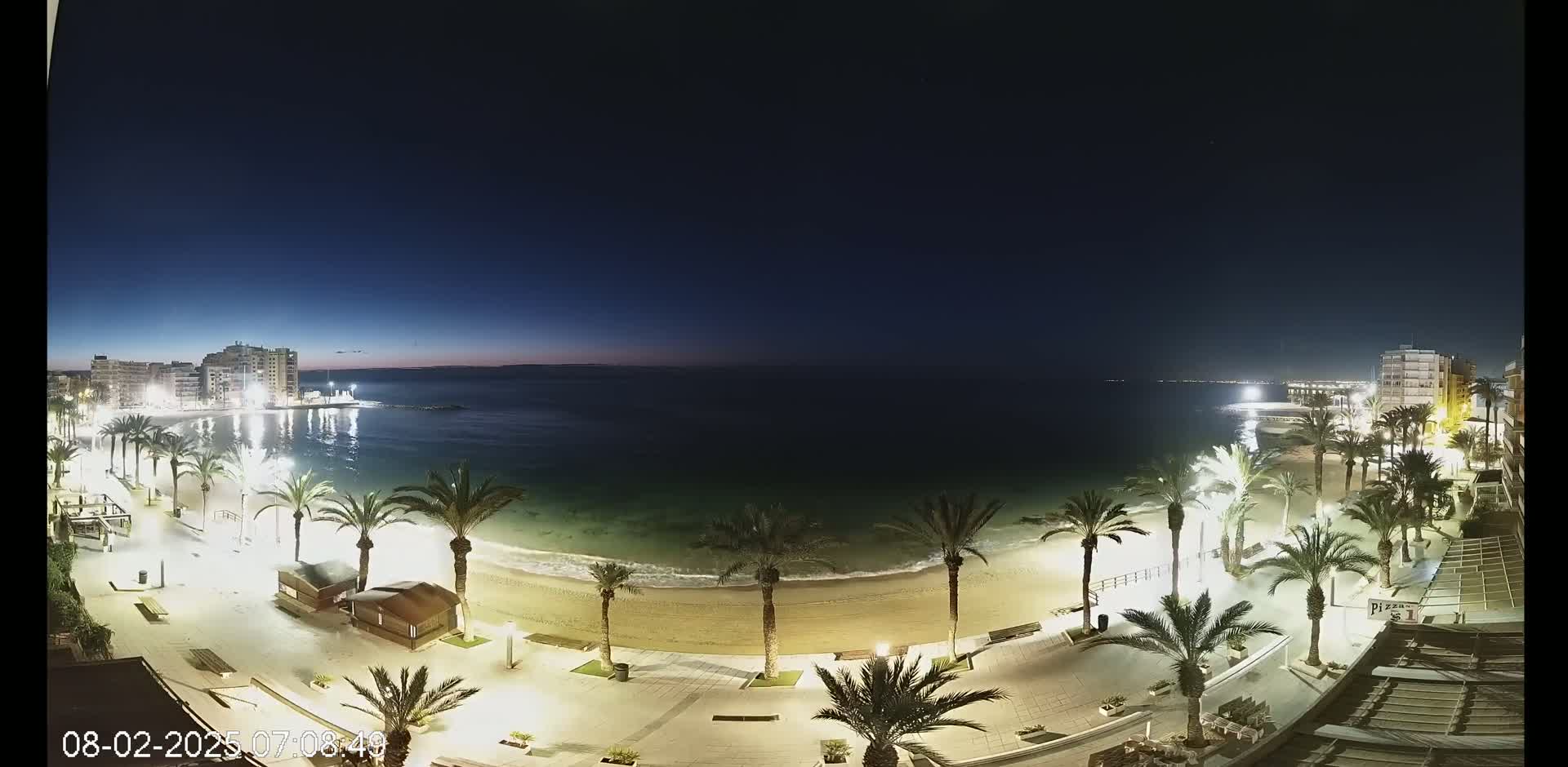 A nighttime beach scene shows a calm sea under a dark blue sky, with palm trees lining a brightly lit promenade next to a sandy beach and buildings in the background.