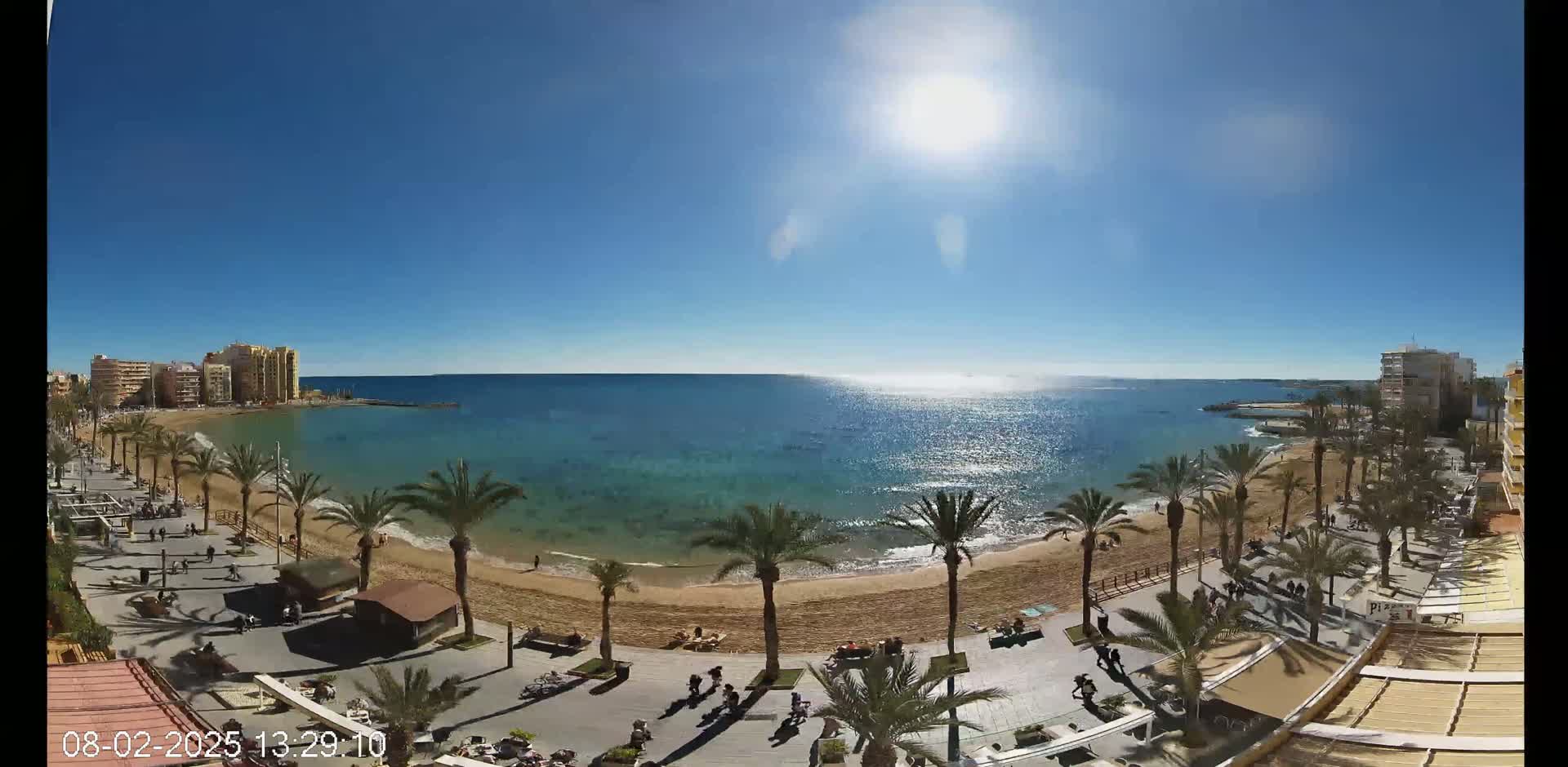 A sunny day shows a sandy beach with palm trees lining a boardwalk, next to buildings overlooking a calm, blue sea.