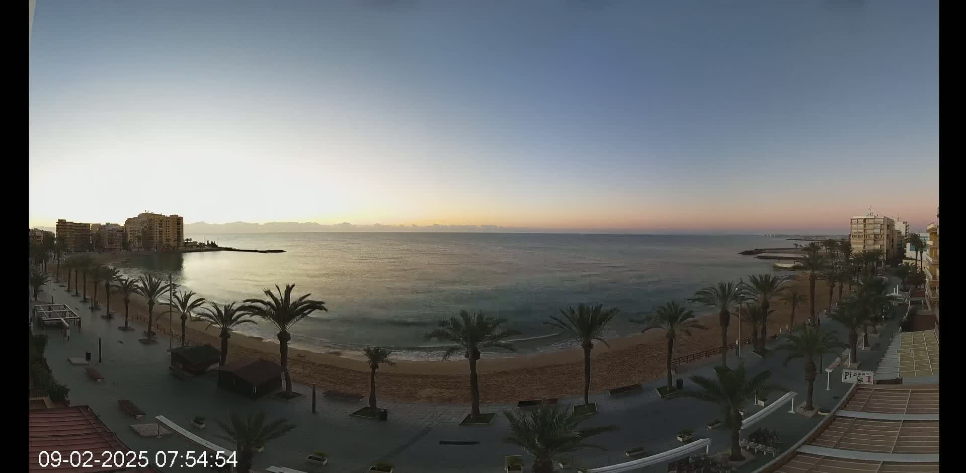 A calm, sunrise-lit ocean beach is lined with palm trees and buildings along a paved walkway.