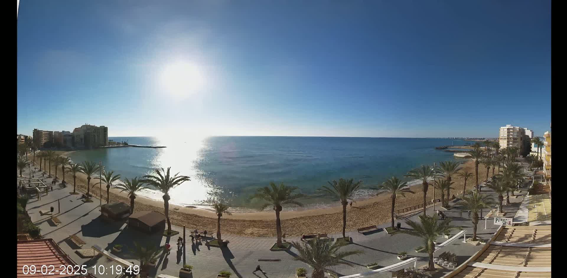 A sunny day at a beach, showing a sandy shore lined with palm trees and buildings, and calm, clear blue water.