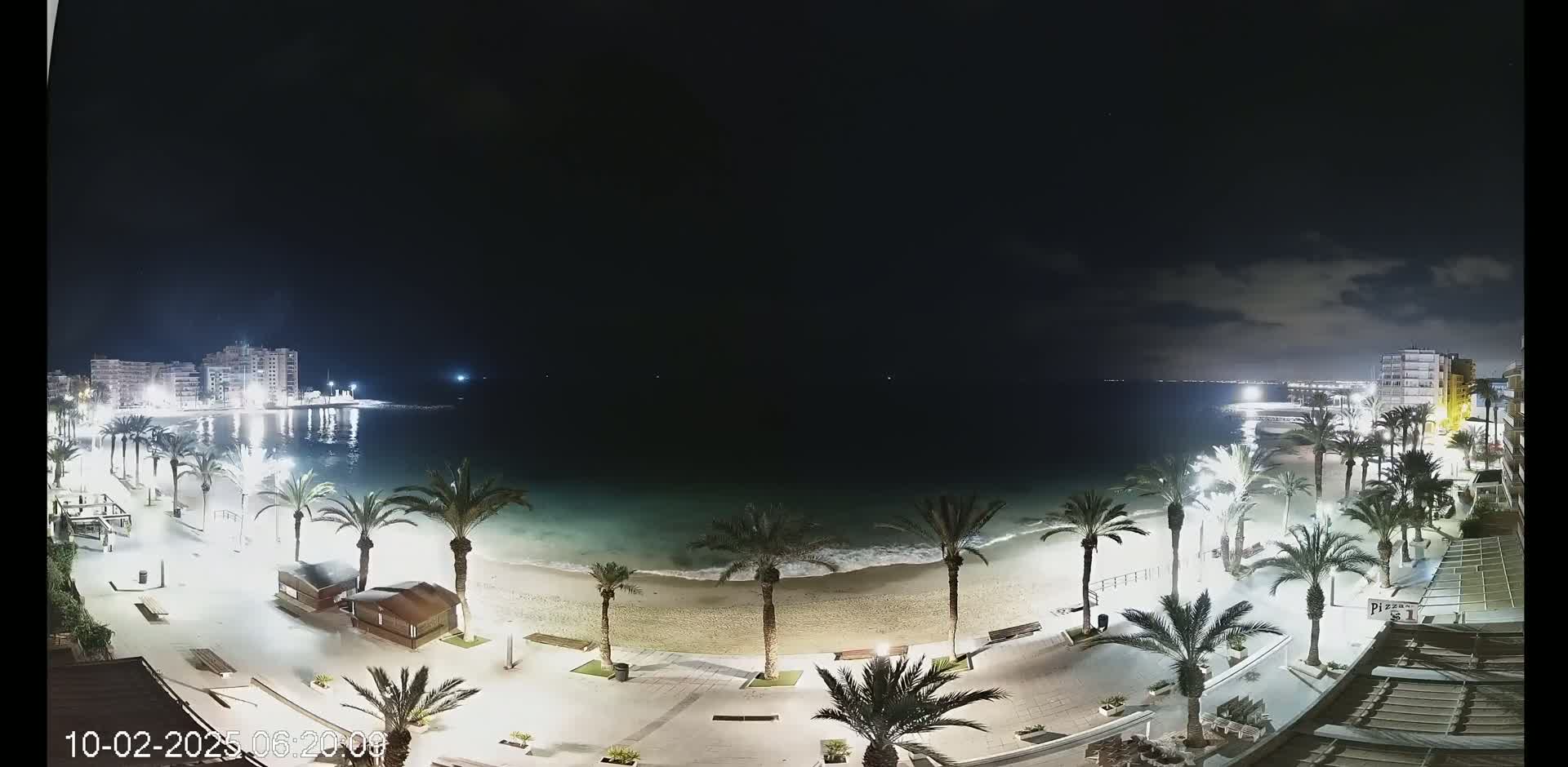 A nighttime view of a beach at the edge of a town, illuminated by streetlights, with palm trees lining the sandy shore and calm waves lapping at the sand.