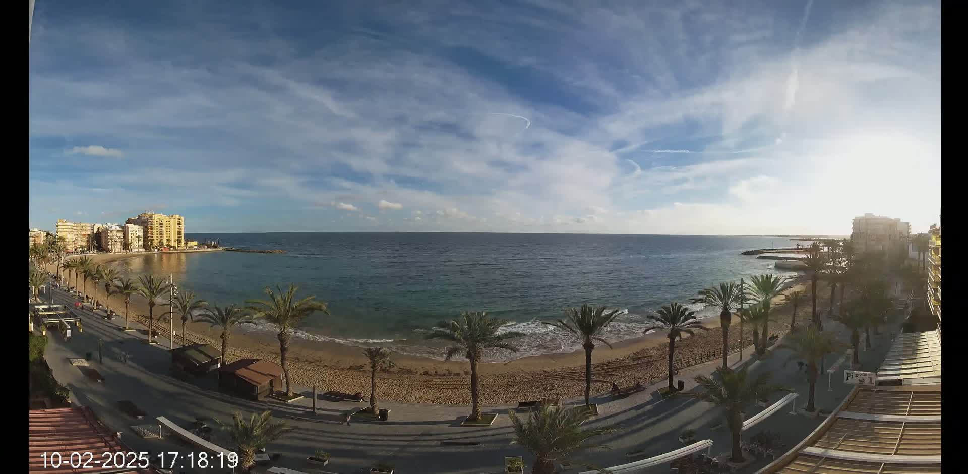 A partly cloudy day shows a sandy beach with palm trees lining a paved walkway, buildings visible along the coast, and a calm ocean in the background.