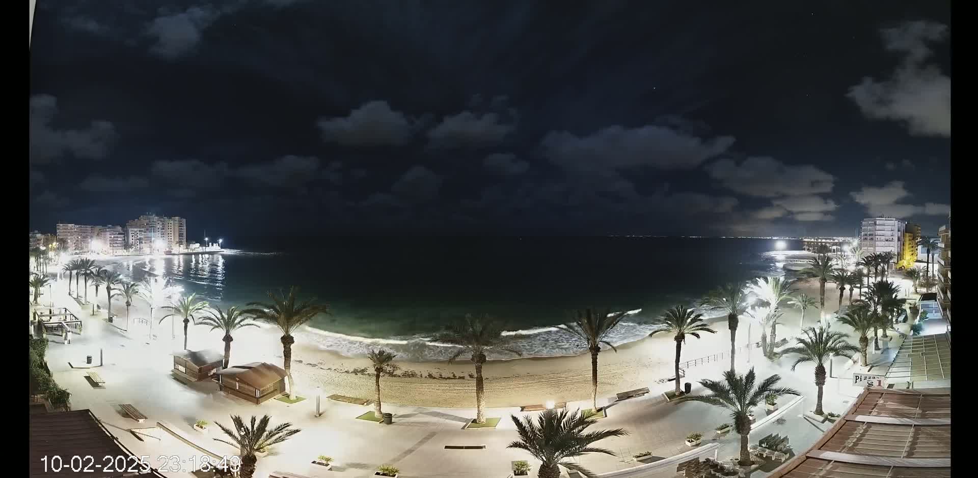 A nighttime view of a beach at the edge of a town, with palm trees lining a paved walkway, gentle waves lapping the shore under a partly cloudy sky.