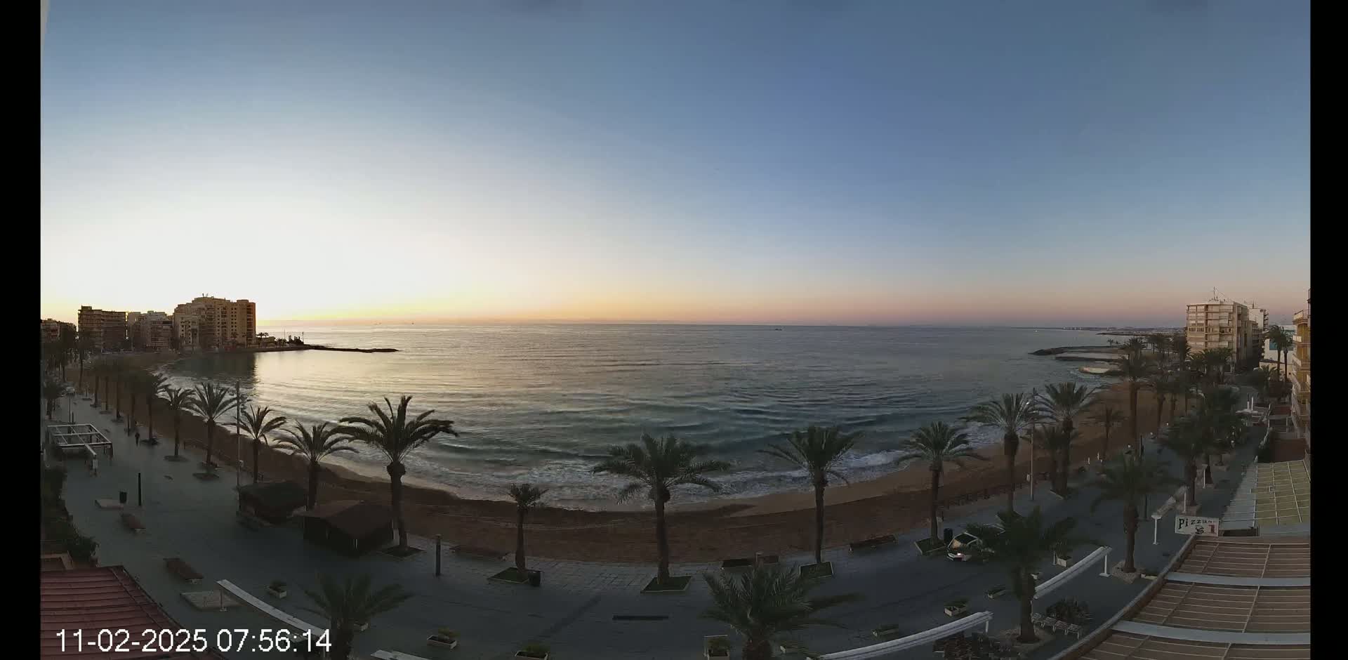 A sunrise illuminates a sandy beach lined with palm trees and buildings, with calm ocean waves gently lapping the shore.