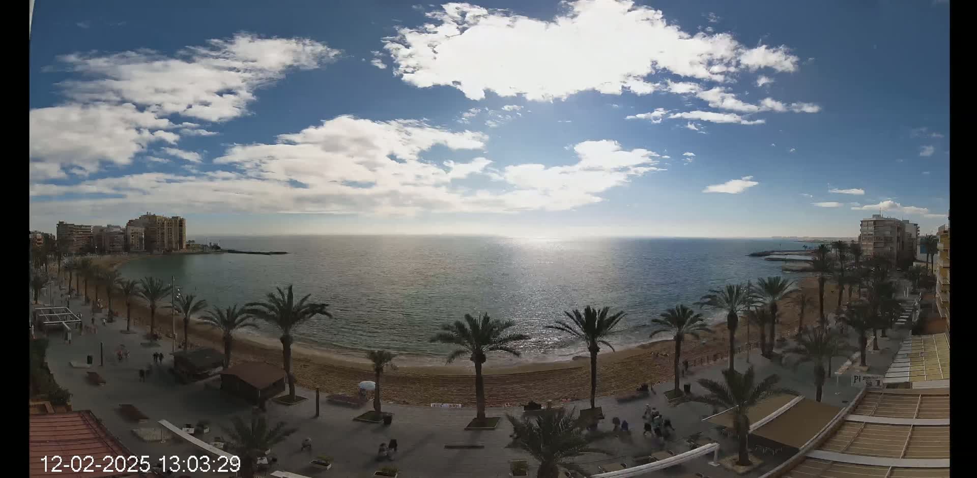 A partly cloudy sunny day overlooks a sandy beach lined with palm trees, buildings, and people walking along a promenade next to a calm sea.