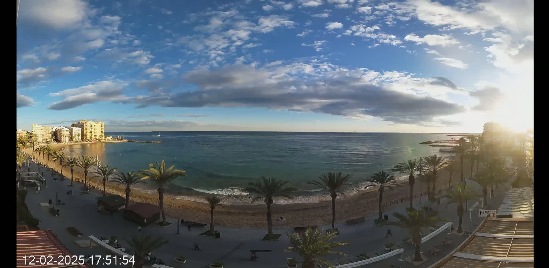 A partly cloudy afternoon shows a beach with palm trees lining a paved walkway next to calm ocean waters and buildings in the distance.