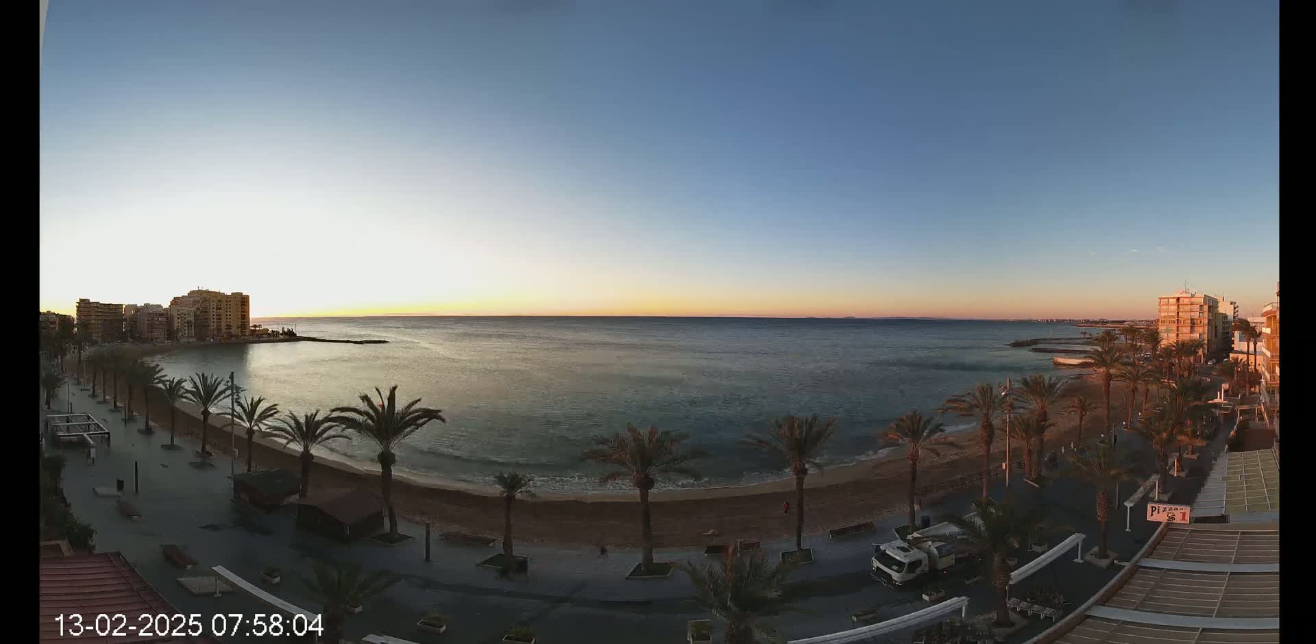 A sandy beach lined with palm trees and buildings stretches along a calm sea at sunrise under a clear sky.