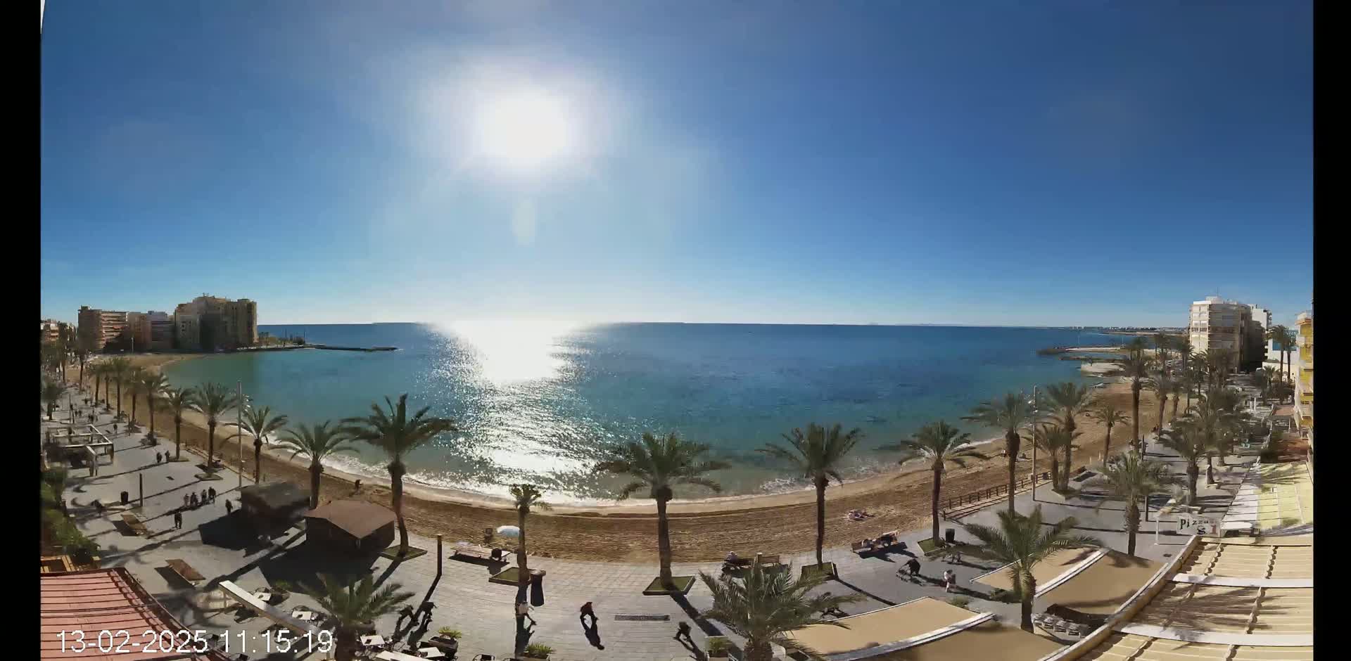 A sunny day shows a sandy beach with palm trees lining a paved walkway, buildings in the background, and a calm blue sea.