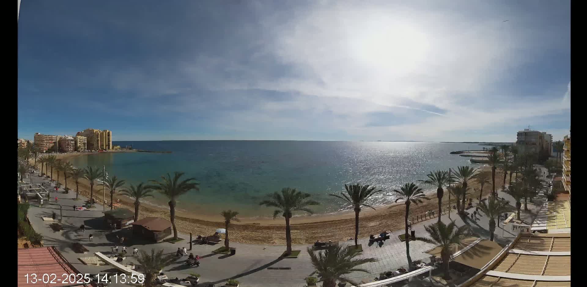 A mostly sunny day shows a sandy beach with palm trees lining a boardwalk and buildings along the coast, overlooking a calm, sparkling sea.