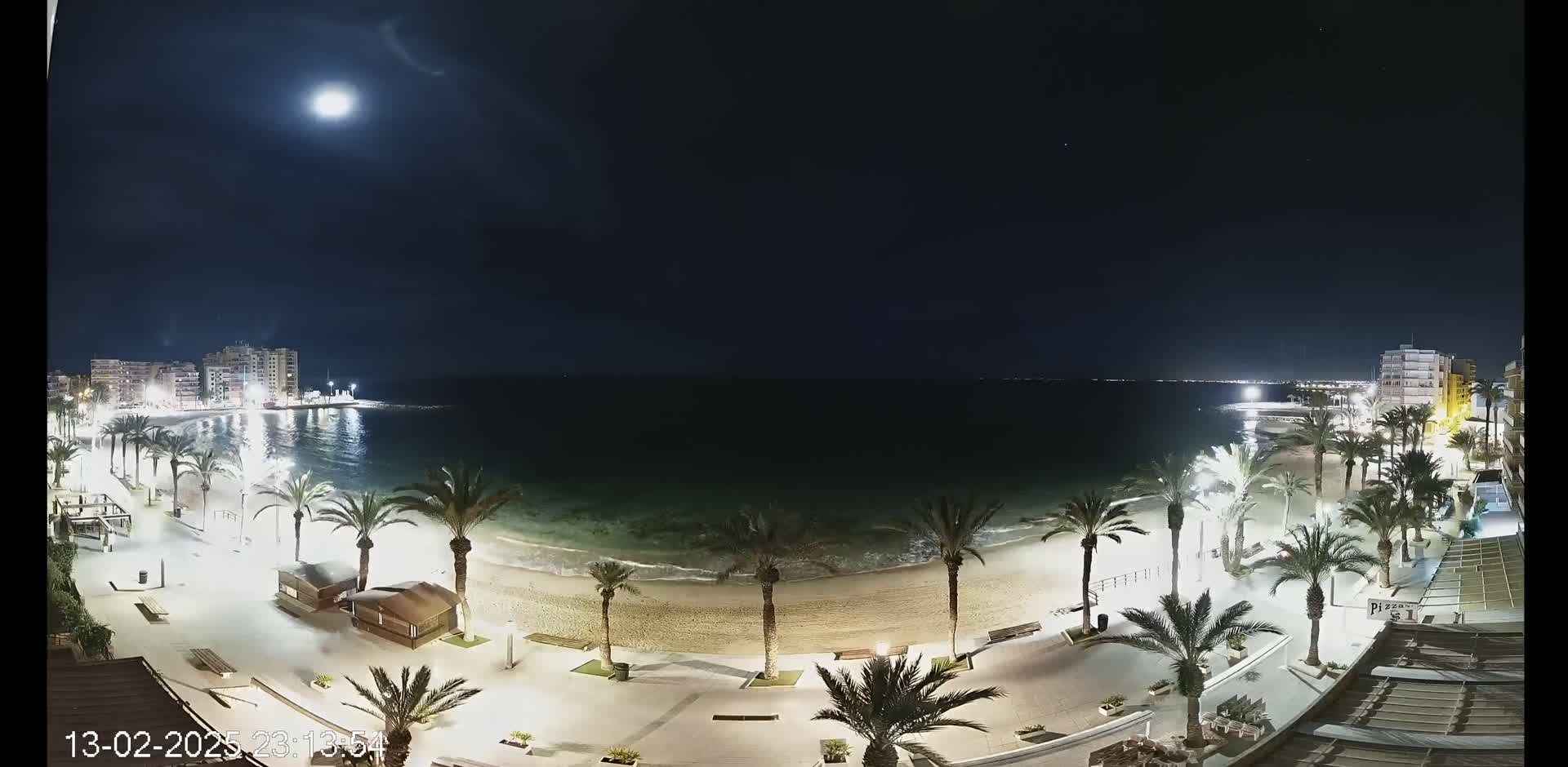 A moonlit night scene shows a sandy beach lined with palm trees and illuminated walkways, with buildings visible in the distance across calm water.