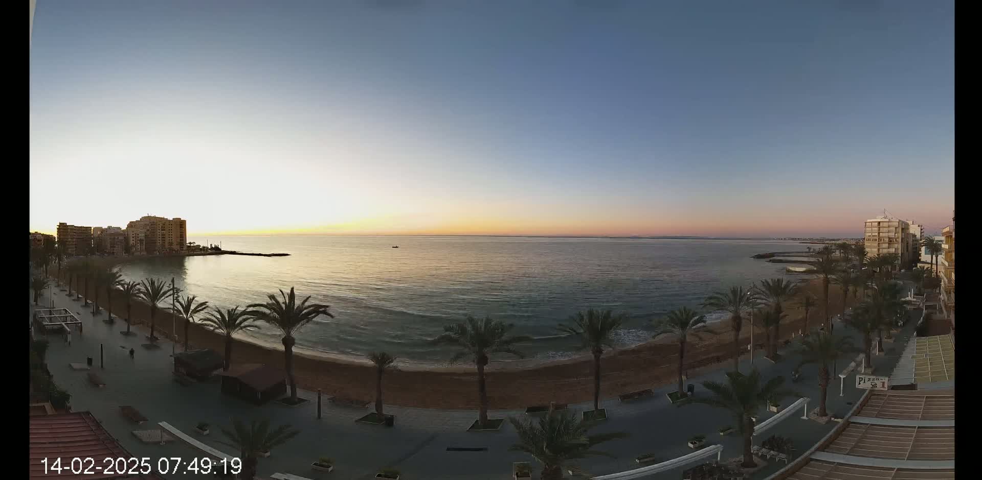 A sunrise over a calm ocean, illuminating a sandy beach lined with palm trees and buildings along a paved walkway.