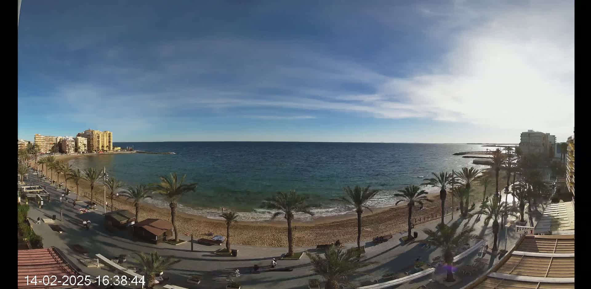 A mostly sunny day at a sandy beach with palm trees lining a promenade, buildings visible in the background, and calm, clear ocean water.