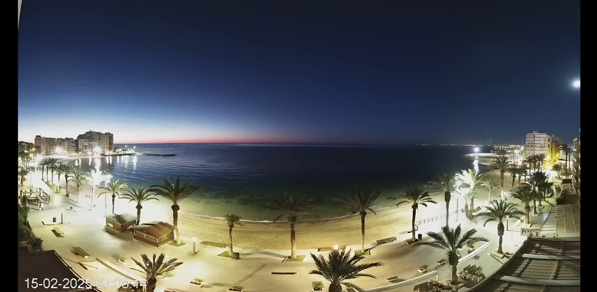 A nighttime, coastal scene shows a sandy beach lined with palm trees and lit walkways, with buildings visible on either side and a calm sea under a dark blue, twilight sky.