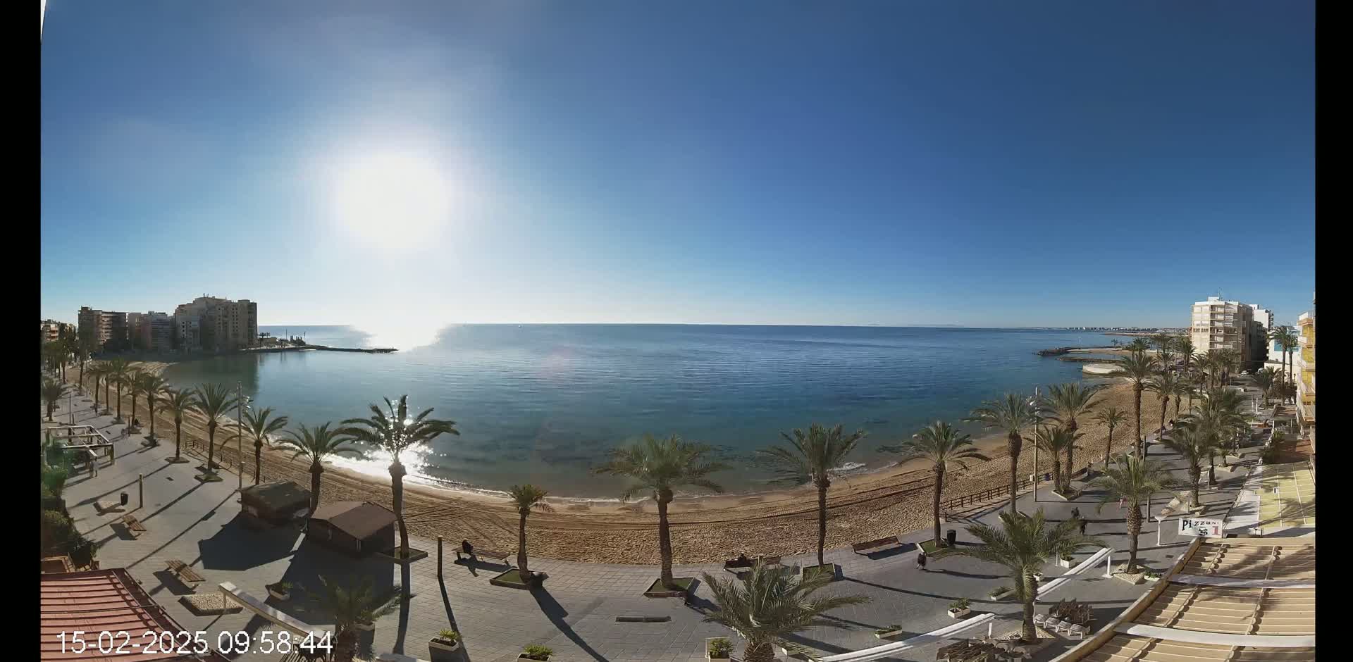 A sandy beach lined with palm trees and buildings stretches along a calm, blue ocean under a bright, sunny sky.