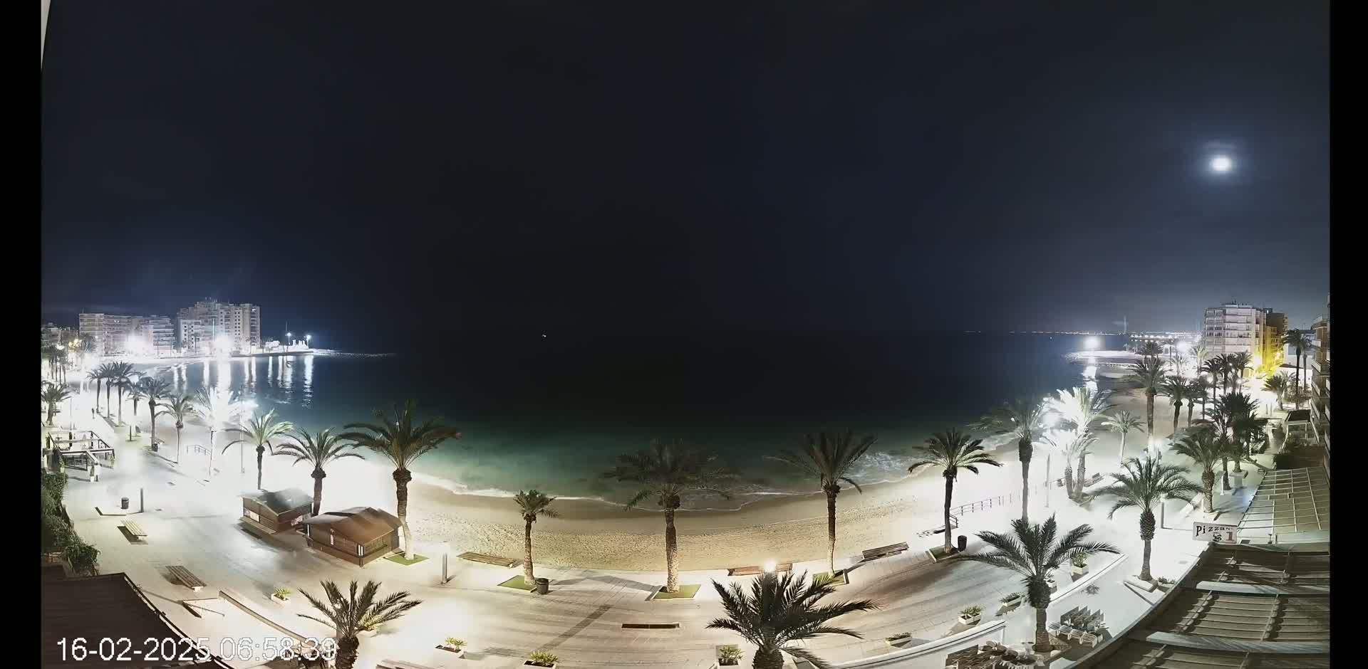 A moonlit nighttime view of a sandy beach lined with palm trees and buildings, with calm water stretching to a distant, illuminated cityscape.