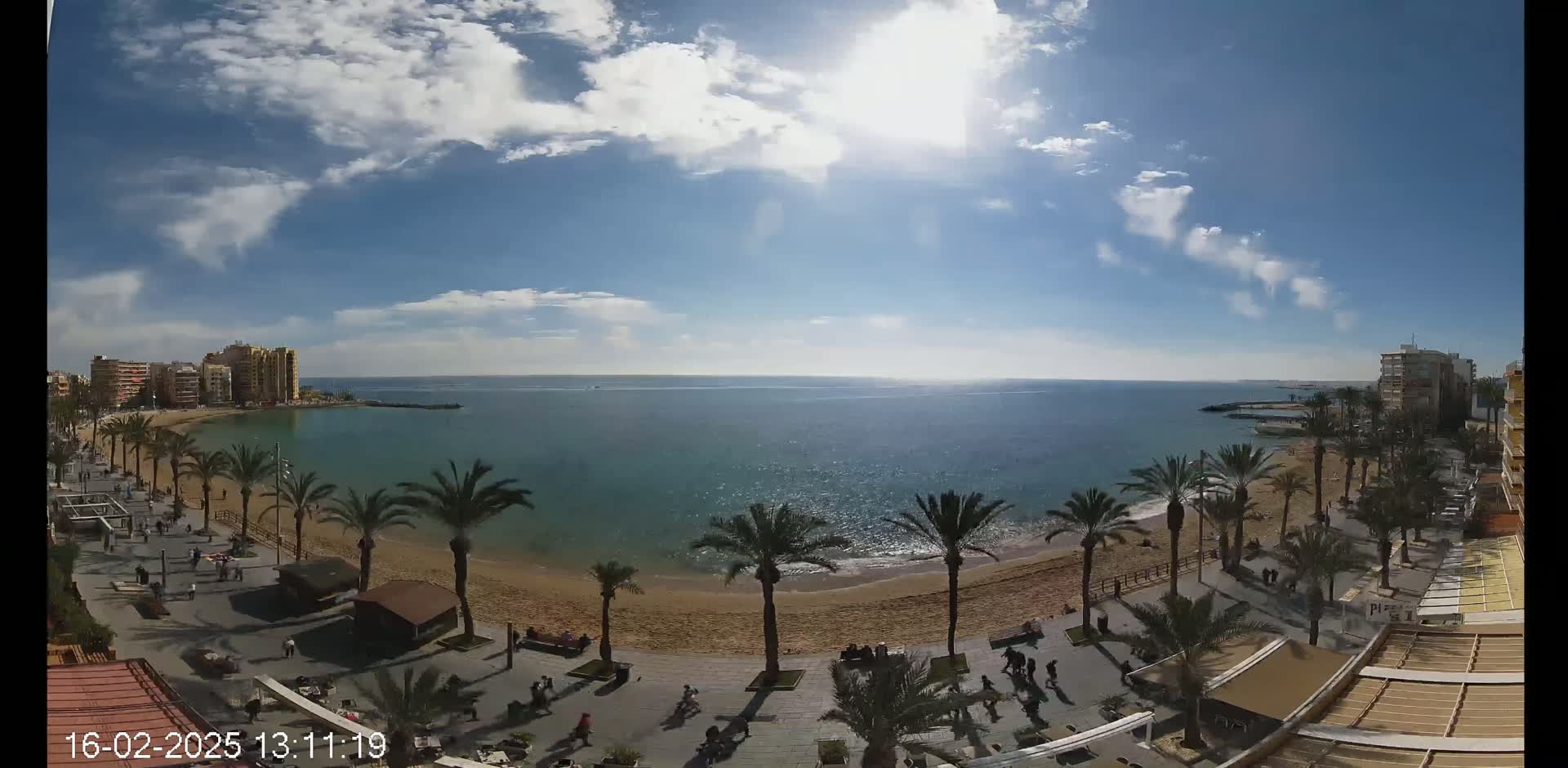 A sunny day with partly cloudy skies shows a sandy beach lined with palm trees and buildings, overlooking a calm blue ocean.