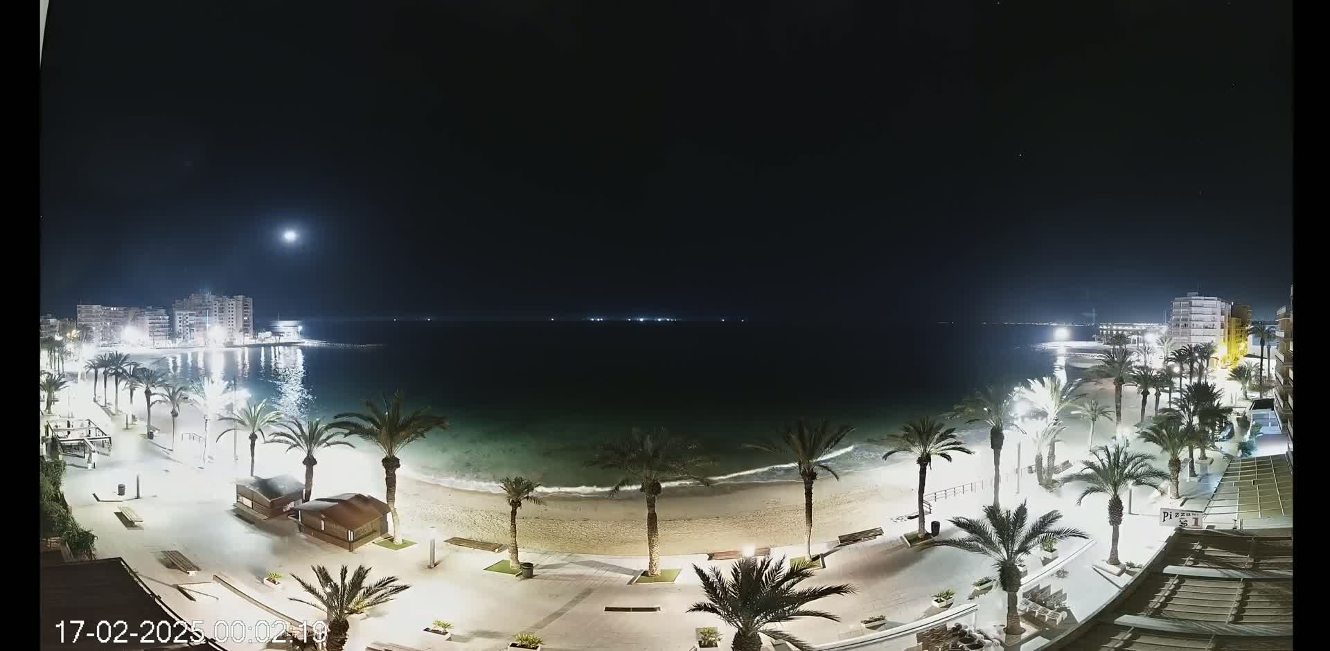 A moonlit night scene shows a sandy beach bordered by palm trees and illuminated buildings, with calm waves lapping the shore.