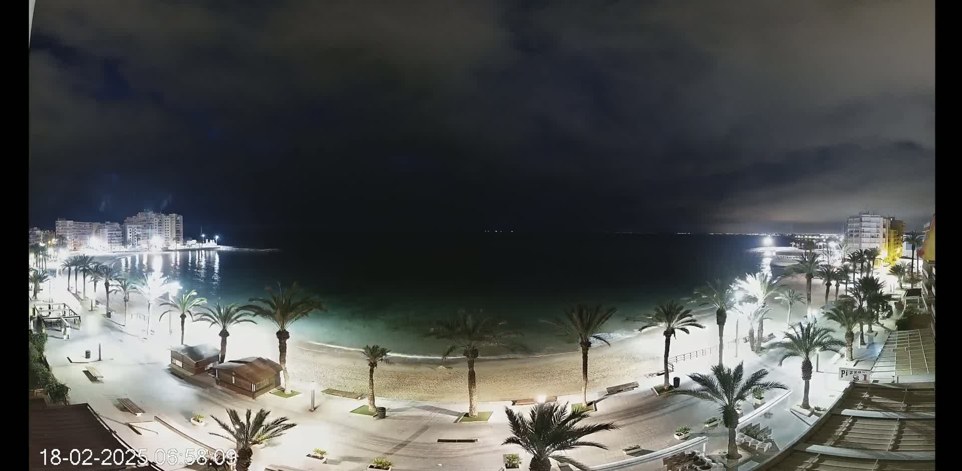 A nighttime view of a beach with palm trees lining a paved walkway, buildings in the distance, and a calm sea under a cloudy sky.