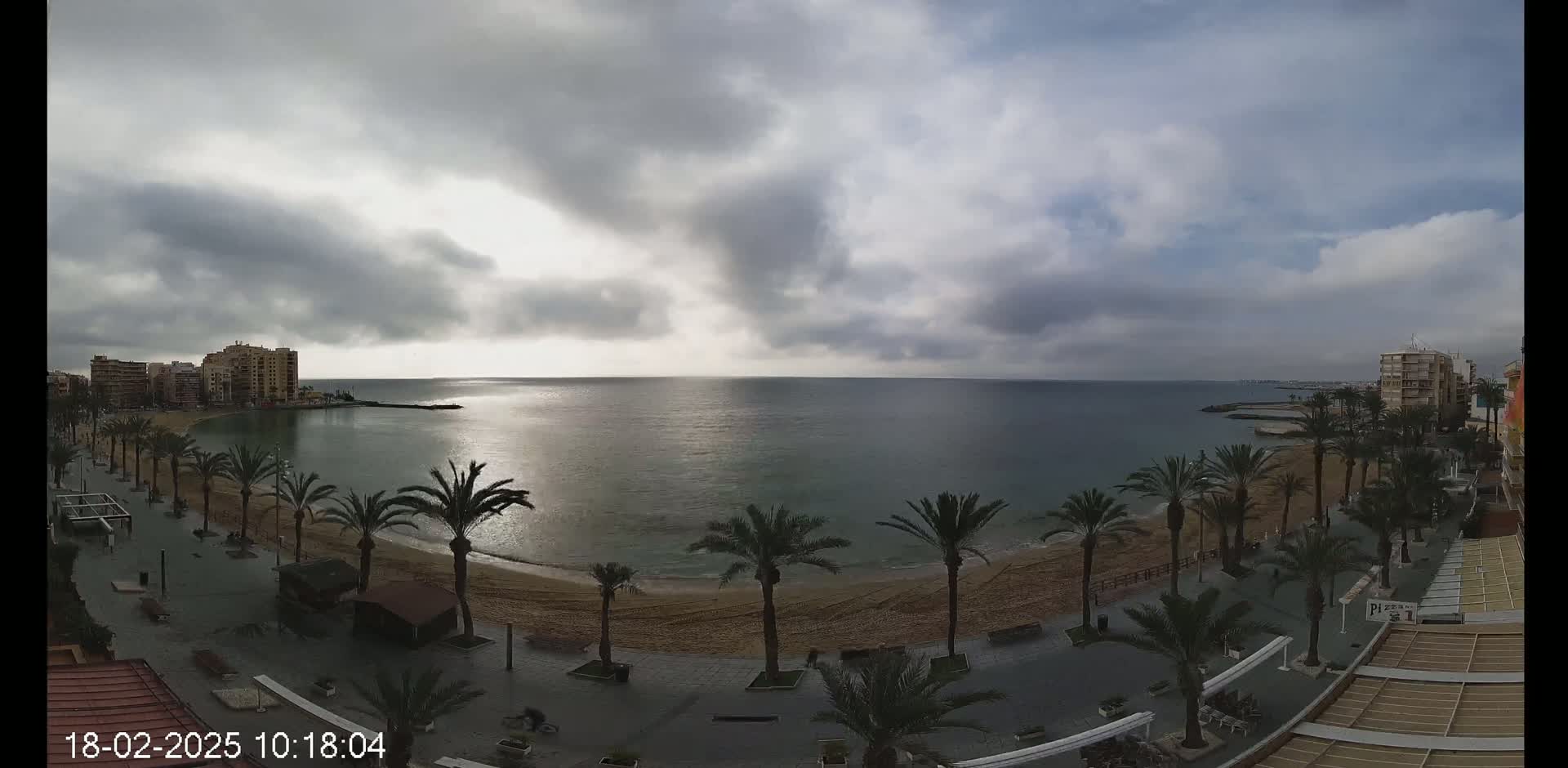 A mostly cloudy sky overlooks a sandy beach lined with palm trees and buildings, with calm ocean waters stretching to the horizon.