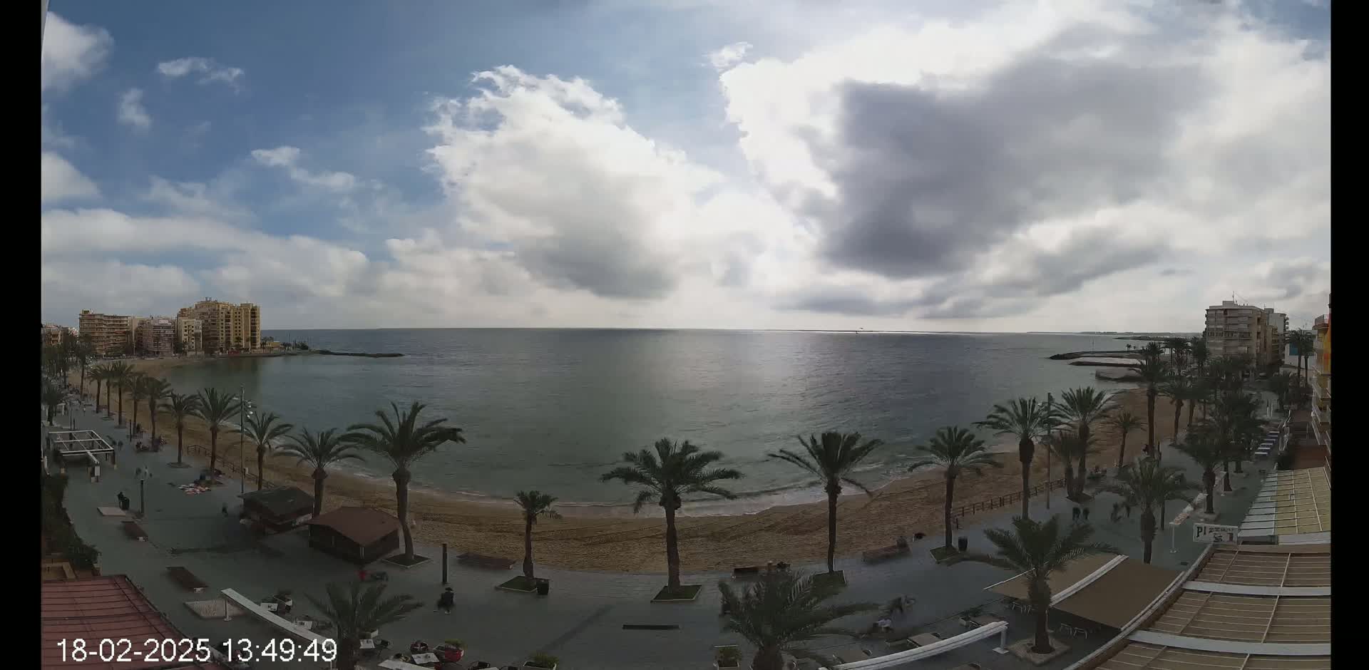 A partly cloudy day overlooks a sandy beach lined with palm trees and buildings along a calm sea.