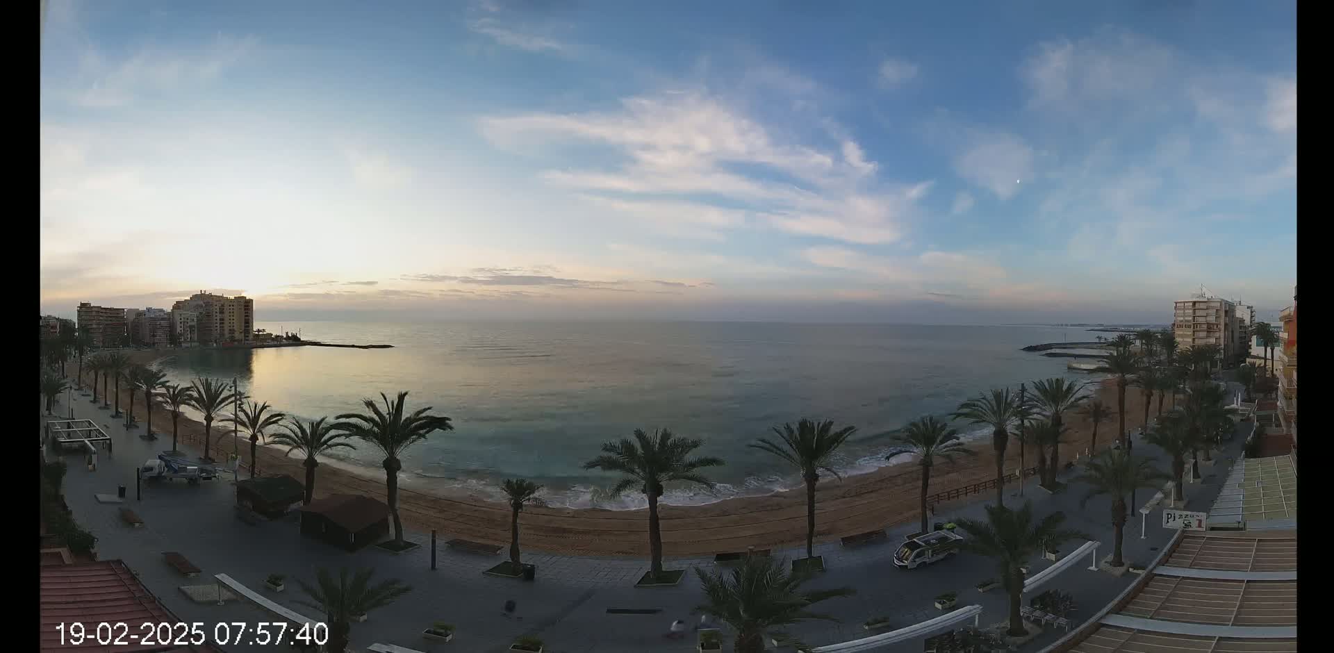 A sandy beach lined with palm trees and buildings sits along a calm ocean under a partly cloudy sky at sunrise.