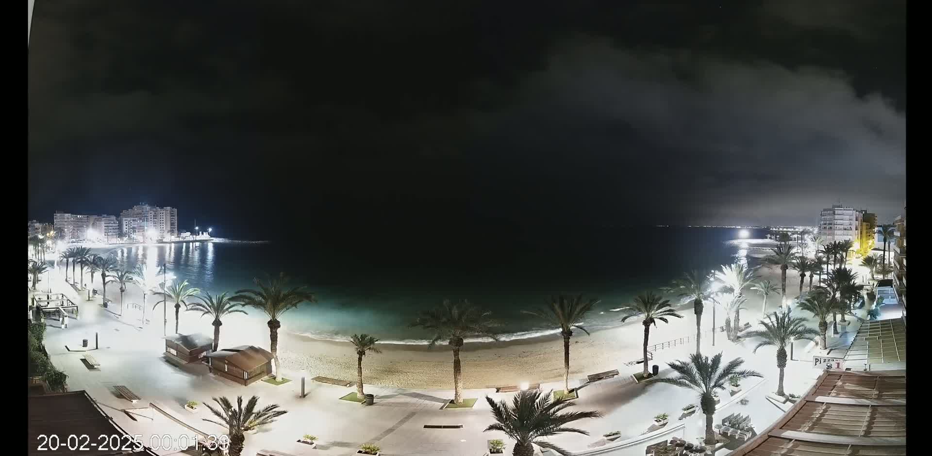 A nighttime view of a sandy beach with palm trees and buildings along a calm ocean under a partly cloudy sky.