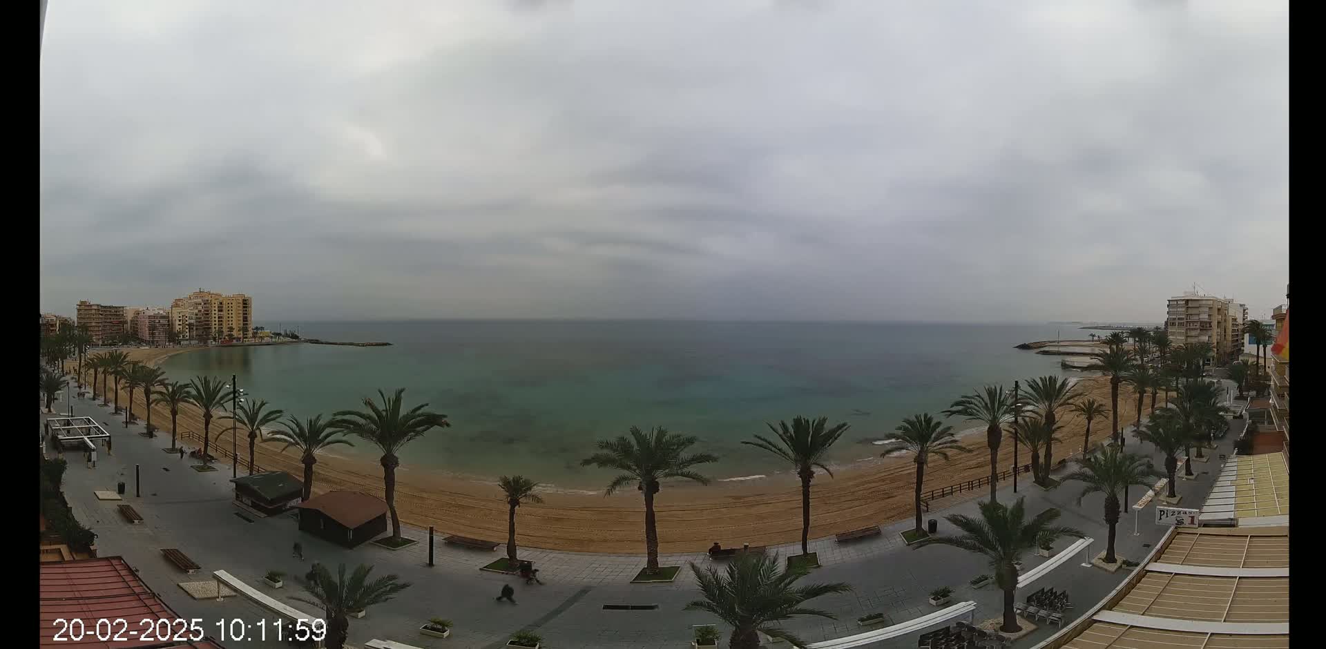 A sandy beach lined with palm trees and bordered by buildings stretches along a calm, teal-colored sea under an overcast sky.
