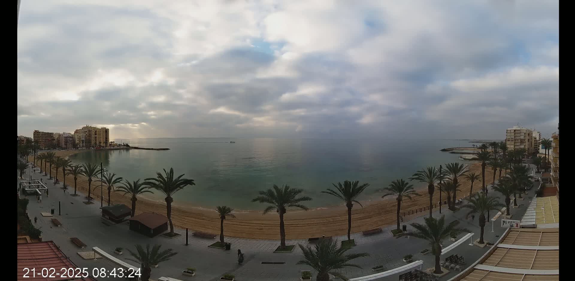A mostly cloudy day reveals a sandy beach lined with palm trees and buildings along a calm, teal-colored sea.