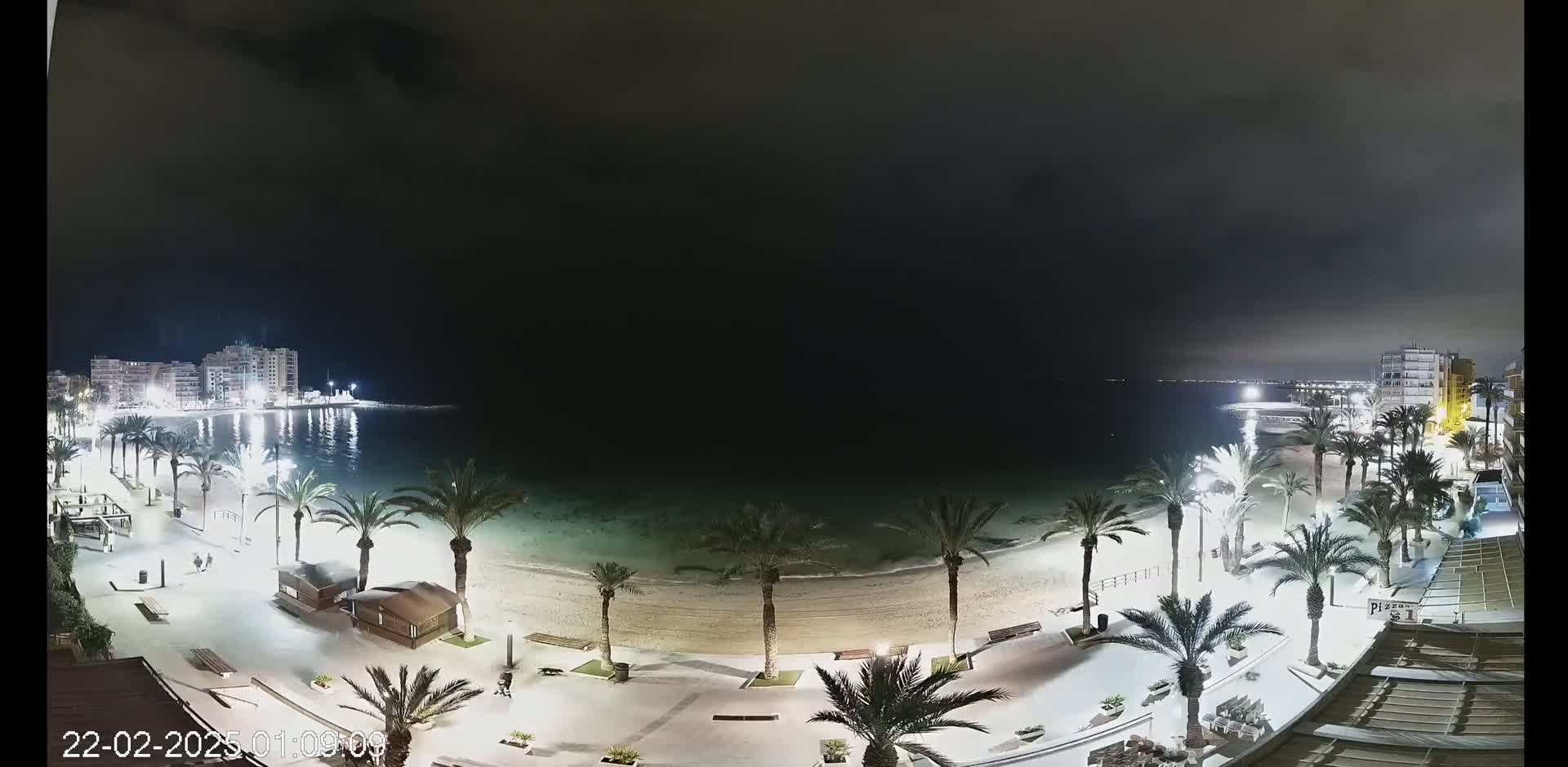 A nighttime view of a sandy beach lined with palm trees and illuminated walkways, overlooking a calm, dark-colored sea with buildings visible in the distance under a mostly cloudy sky.