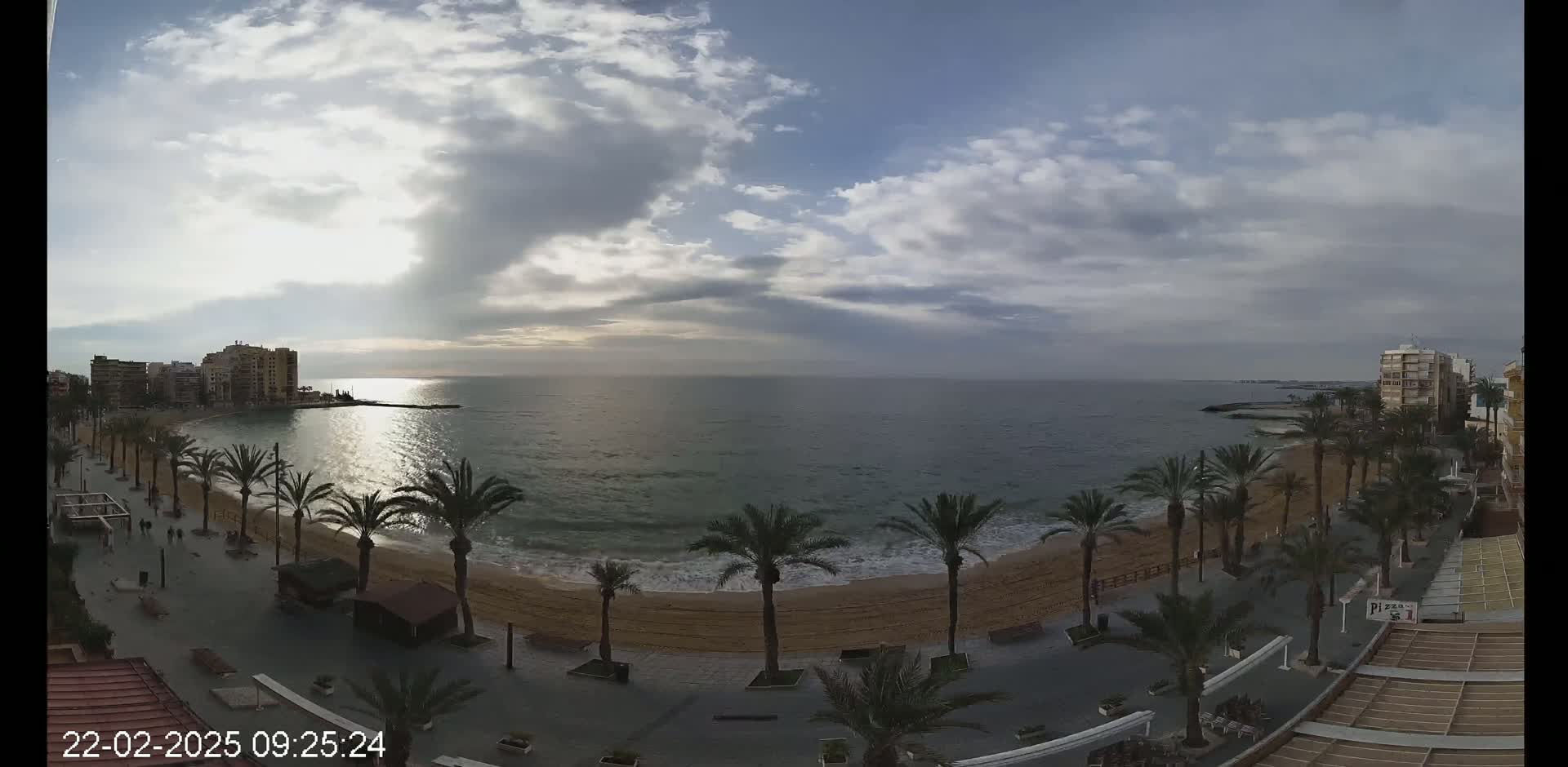 A partly cloudy day shows a sandy beach lined with palm trees and buildings, overlooking a calm sea.
