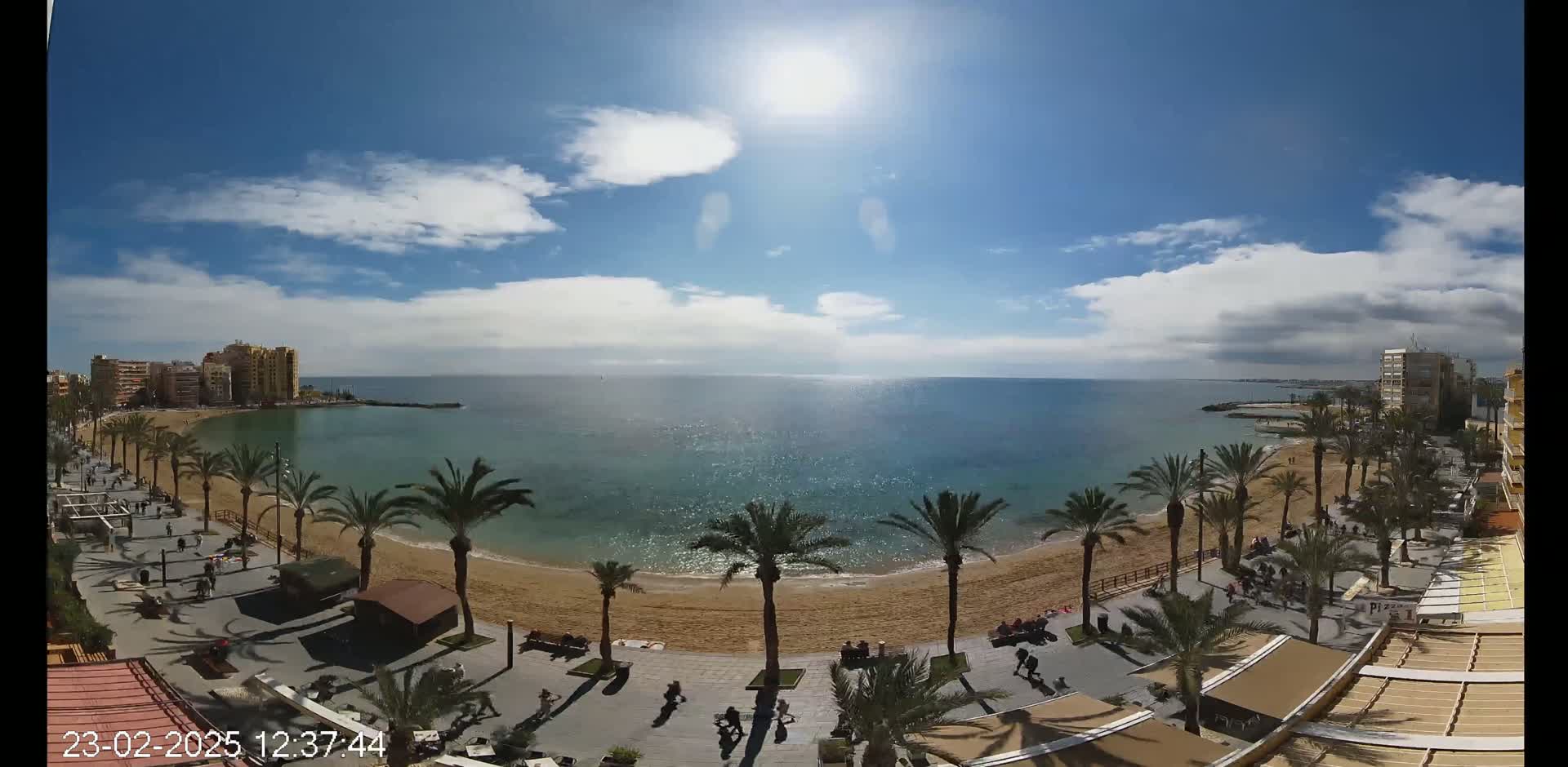 A sunny day with partly cloudy skies shows a sandy beach lined with palm trees and bordered by buildings, a paved walkway, and people strolling along the shore.