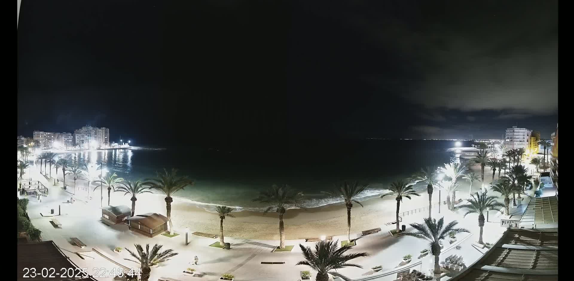 A nighttime beach scene shows a sandy shore lined with palm trees and buildings under a partly cloudy sky.