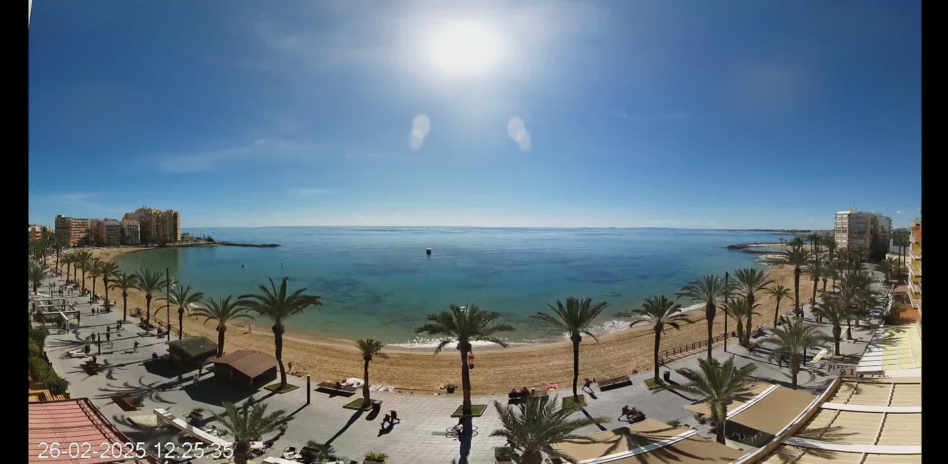 A sunny day shows a sandy beach lined with palm trees and buildings, with calm blue water extending to a distant horizon.