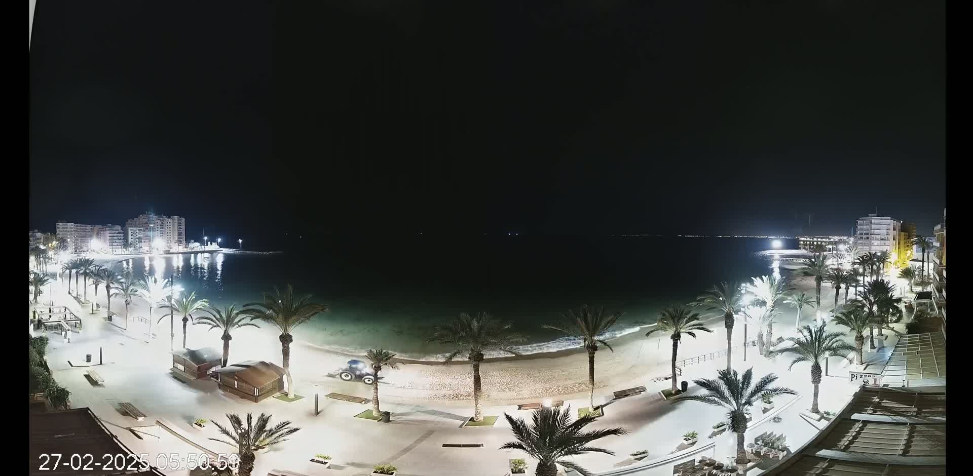 A nighttime beach scene shows a sandy shore lined with palm trees and illuminated walkways, buildings in the background, and a calm, dark sea under a clear sky.
