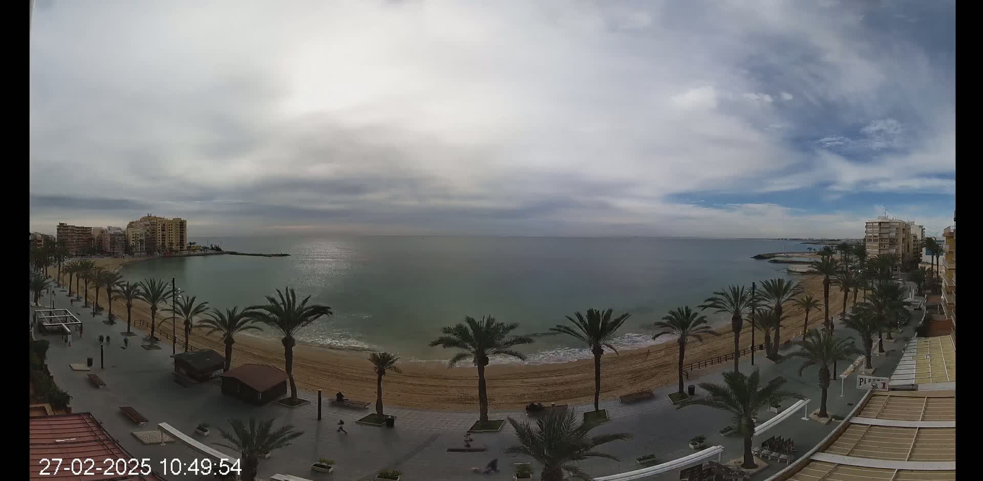 A mostly cloudy day reveals a sandy beach lined with palm trees and buildings along a calm sea.