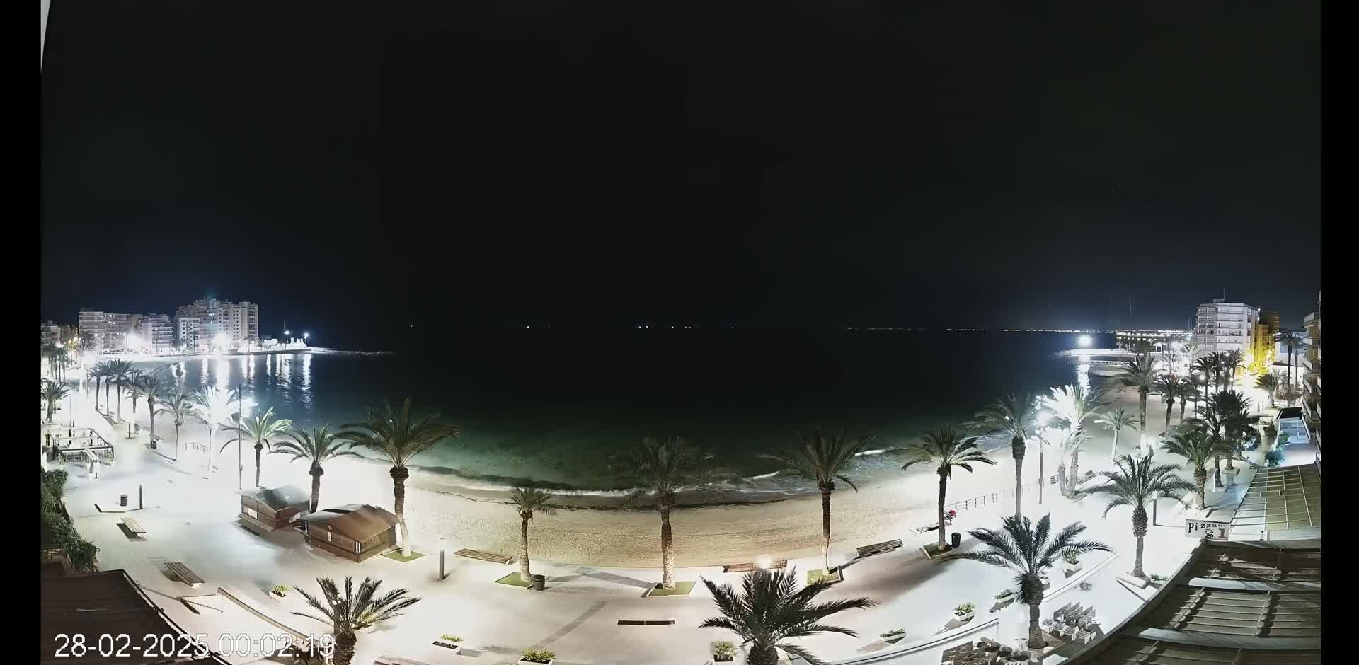 A nighttime beach scene shows a sandy shore lined with palm trees and illuminated walkways, extending to buildings along the calm, dark water.