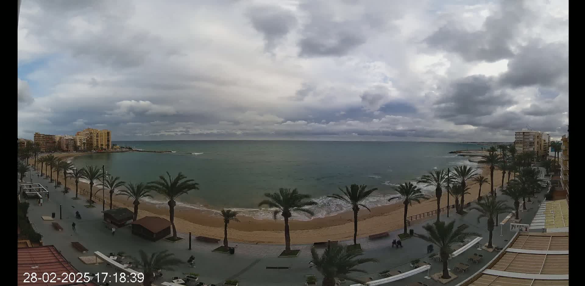 A beach with palm trees lining a promenade, overlooks a calm sea under a cloudy sky.